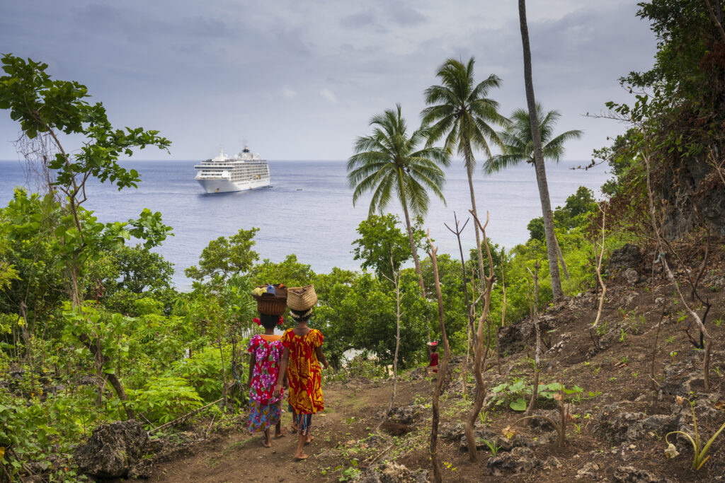 natives walking with baskets on head with cruise ship in the ocean in background